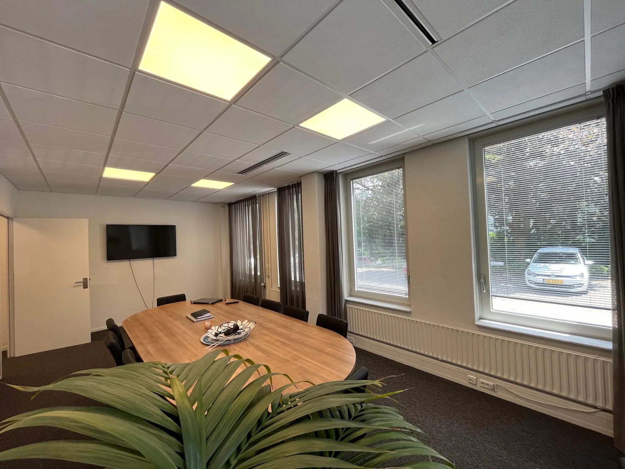 Vergaderruimte aan de Oude Utrechtseweg met ovale houten tafel, stoelen, groot beeldscherm en uitzicht op geparkeerde auto’s buiten.